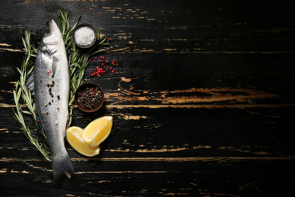 Fresh uncooked sea bass fish, lemon and spices on black wooden background - Photo, Image