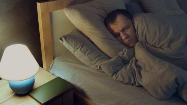 Mężczyzna leży w łóżku w nocy, pokryty kocem, nie może spać, martwić się, patrzy na lampę - Materiał filmowy, wideo