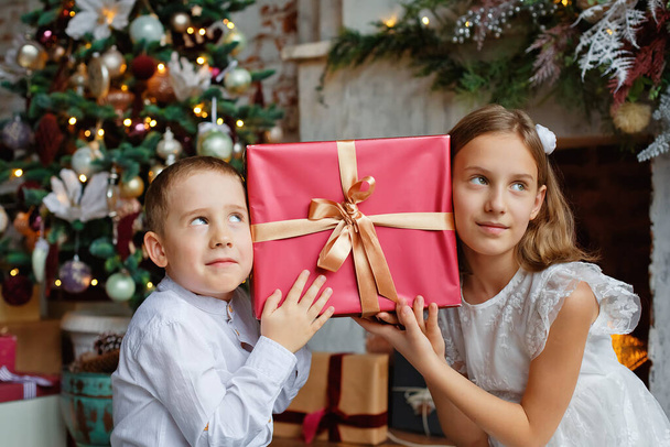 Τα παιδιά έσκυψαν τα κεφάλια τους στο δώρο. Ένα κορίτσι και ένα αγόρι με δώρα κάτω από το χριστουγεννιάτικο δέντρο. Αδελφός και αδελφή είναι ευχαριστημένοι με τα δώρα της Πρωτοχρονιάς. - Φωτογραφία, εικόνα