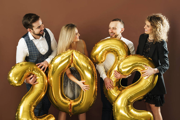 2022 yılını kutlayan bir grup güzel giyinmiş parti insanı kahverengi arka planda altın bir balonla - Fotoğraf, Görsel