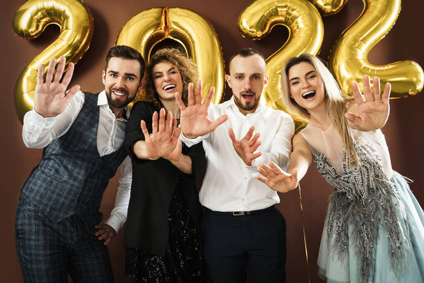 Group of beautiful well dressed party people celebrating arrival of 2022 New Year with a golden balloons on brown background - Φωτογραφία, εικόνα