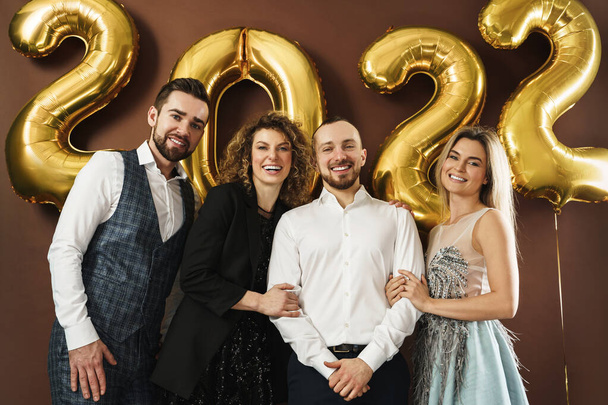 Group of beautiful well dressed party people celebrating arrival of 2022 New Year with a golden balloons on brown background - Photo, image