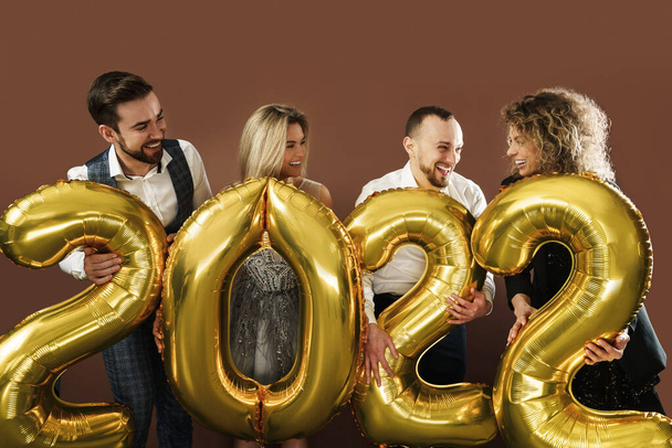 Group of beautiful well dressed party people celebrating arrival of 2022 New Year with a golden balloons on brown background - Foto, Imagen