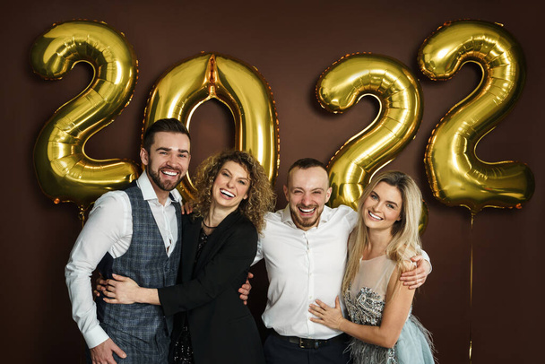 Groep van mooie goed geklede party mensen vieren aankomst van 2022 Nieuwjaar met een gouden ballonnen op bruine achtergrond - Foto, afbeelding
