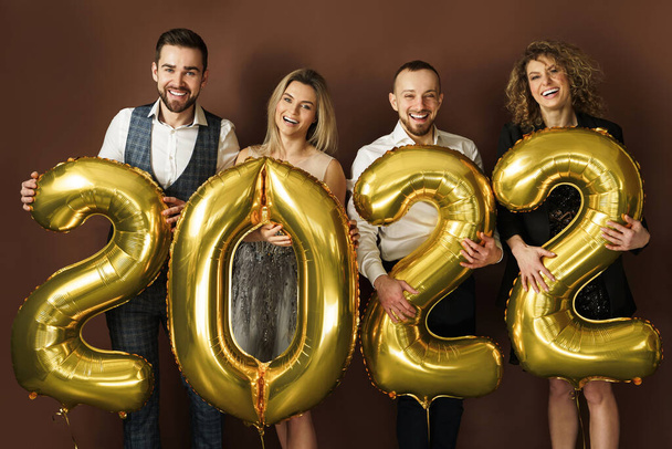 Gruppe schön gekleideter Partygänger feiert die Ankunft des neuen Jahres 2022 mit goldenen Luftballons auf braunem Hintergrund - Foto, Bild
