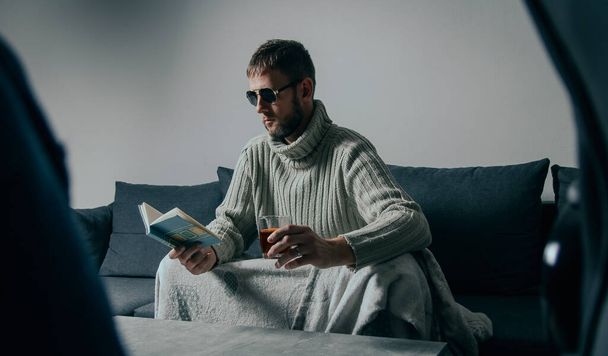 Portrait of young caucasian man in sunglasses sitting on couche reading book and drinking alcohol. Cinematic people concept - 写真・画像