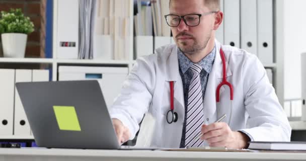 Fiatal orvos szemüveggel nézi laptop képernyő és írás orvosi dokumentumok 4k film - Felvétel, videó