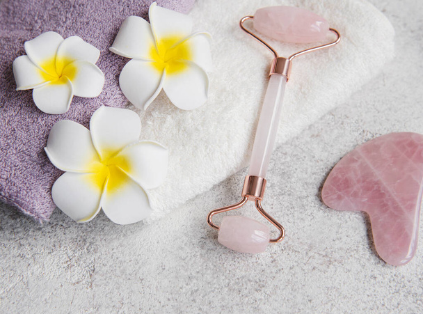 Beautiful spa relax concept. Towel, jade roller and frangipani flowers on grey stone background - Photo, Image