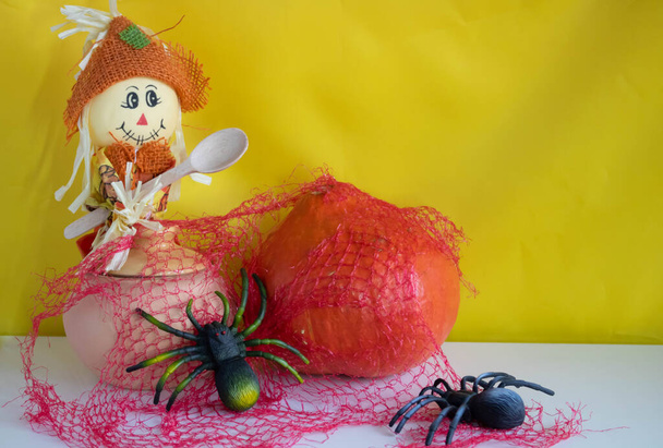 Een vogelverschrikker met een houten lepel, een kleipot, een pompoen en twee spinnen met een rood web op een witte achtergrond.Halloween en Thanksgiving Day Concept. - Foto, afbeelding