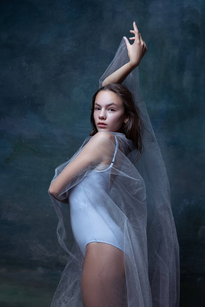 Close-up of young beautiful girl, ballerina posing with white transparent cloth isolated on dark vintage studio background. - Photo, image