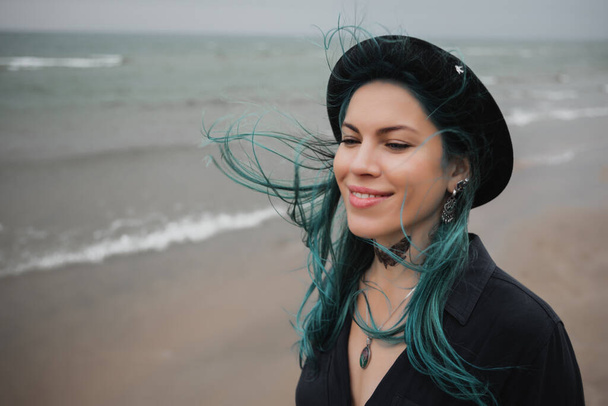 beautiful woman with blue hair and hat walks on the windy beach - Foto, imagen