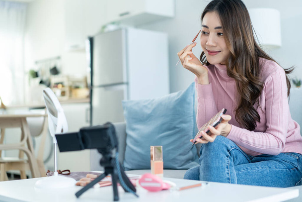 Asian female vlogger talk with camera to review the cosmetic at home. Beautiful Beauty blogger Influencer girl sit on sofa and live streaming, showing eyeshadow palettes makeup to followers in house. - 写真・画像
