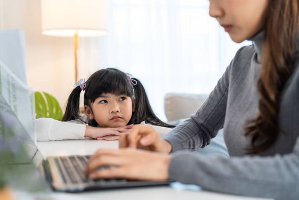 Asian young kid daughter feeling sad and need to play with busy mother. Attractive beautiful mom use laptop computer working from home during new normal and have no time for little child girl in house - Foto, Imagem