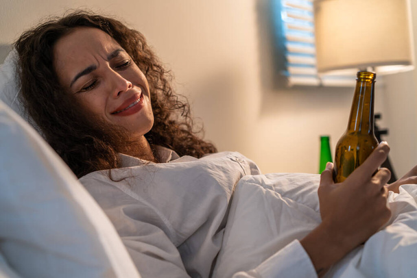 Drunk Latino woman hold beer bottle, and crying feel hangover on bed. Young beautiful depression female lying and crying in tears in bedroom, feel heart broken and celebrate life with alcohol at night - Photo, Image
