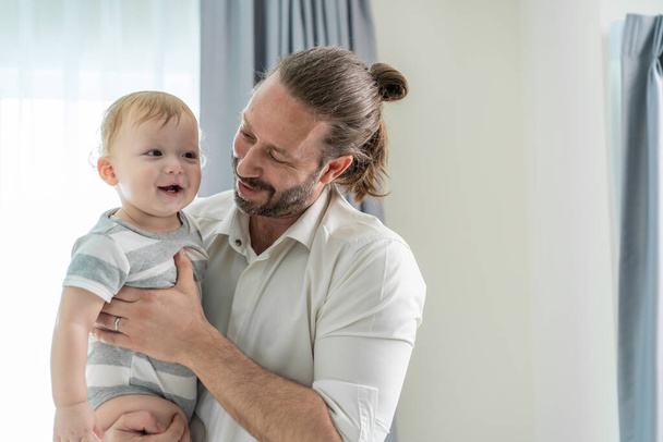 Beyaz bir baba, erkek bebeğini oturma odasında tutuyor. Mutlu aile, çekici genç baba küçük oğlunu elinde mutlulukla evde taşıyor. Evde etkinlik eğlencesi ilişkisi. - Fotoğraf, Görsel