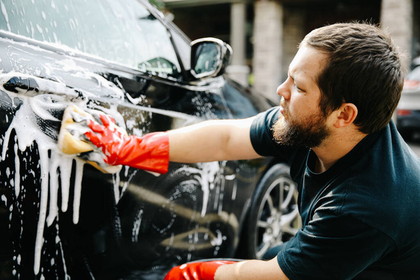 Close up caucasian man washing own black car outside with sponge, foam and wearing red gloves. - Photo, Image