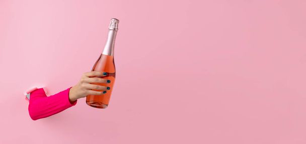 Γυναικείο χέρι με μπουκάλι ροζέ κρασί σπάει μέσα από ροζ χάρτινο φόντο. - Φωτογραφία, εικόνα