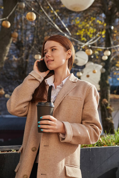 femme caucasienne en manteau beige élégant appelant par smartphone, boire du café - Photo, image
