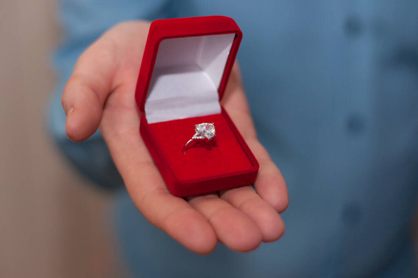 A white gold ring with a precious stone in an open red velvet box in the hand of a man in a blue shirt - Fotoğraf, Görsel