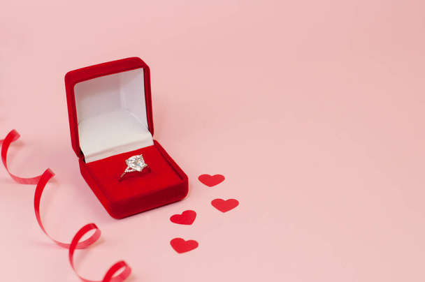 Red velvet box with a white gold ring with a precious stone on a pink background with confetti of red hearts and a red serpentine - Фото, изображение