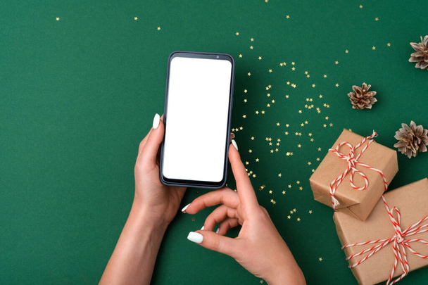 Çağdaş akıllı telefonu olan, yeşil arka planı olan ve Noel süslemeli kadın eller. Düz şablon. Firavun çelengi, hediye kutusu. - Fotoğraf, Görsel