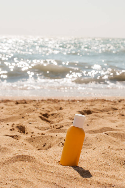 A yellow jar with a white cap with sunscreen stands in the sand on a yellow sandy beach against the background of a blue sea in a vertical format - Fotó, kép