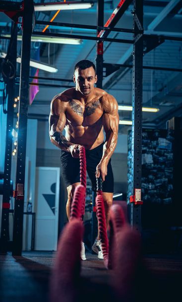 Front view of young man, muscled athlete, bodybuilder training alone at sport gym, indoors. Concept of sport, activity, healthy lifestyle - Valokuva, kuva