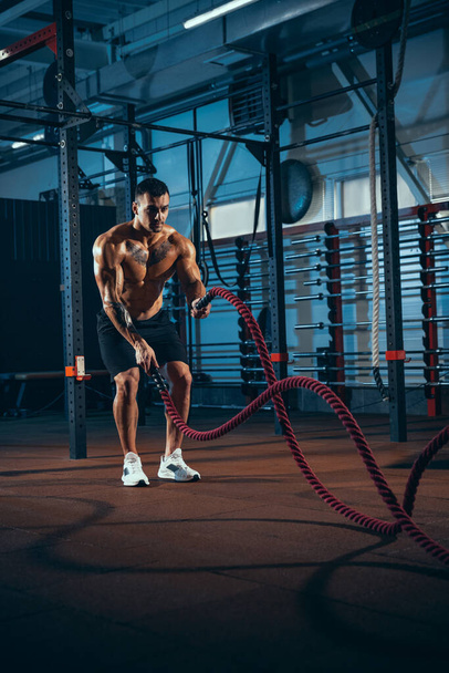 Front view of young man, muscled athlete, bodybuilder training alone at sport gym, indoors. Concept of sport, activity, healthy lifestyle - 写真・画像