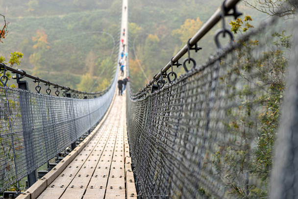 Suspension wooden bridge with steel ropes over a dense forest in West Germany, visible tourists on the bridge in misty weather. - Photo, Image