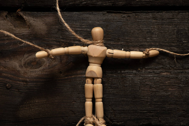 wooden man on ropes on a wooden background, tied to a man's hand, manipulation of people and slavery, free will, power over people - Photo, Image