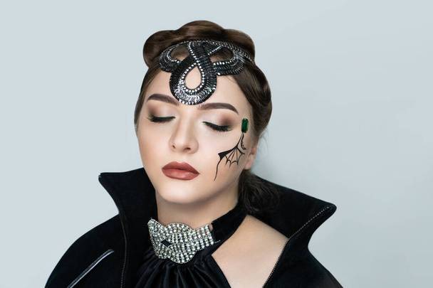 Beautiful makeup for a glamorous Halloween party. Face art on the white skin of the model. Bat wing, snake decoration on the head, silver jewellery. Long black raincoat with stand-up collar. Make-up - Fotó, kép