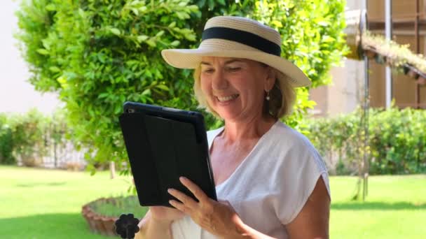 sonriente Senior mujer rubia mujer de negocios en sombrero utiliza tableta PC, tableta digital para el trabajo de negocios o estudio en su propio jardín verde. mujer de 50 a 55 años. El concepto de su propia empresa y formación - Imágenes, Vídeo