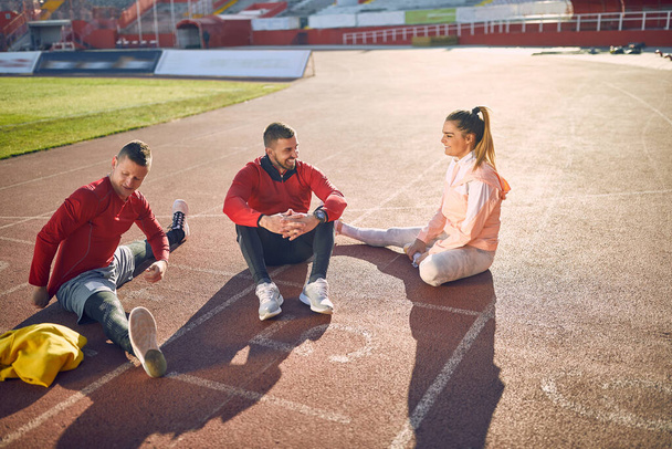 young adult caucasian beardy guy sitting on athlete track together with smiling couple to whom is talking. personal trainer concept - Φωτογραφία, εικόνα