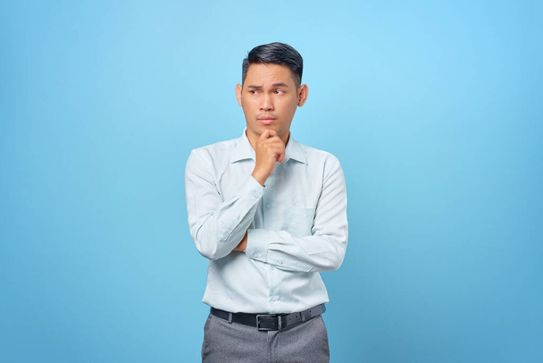 Pensive young handsome businessman rubs chin and thinking about a question on blue background - Фото, изображение