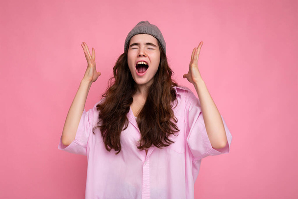 Красива емоційна щаслива молода кучерява брюнетка в рожевій сорочці та сірому капелюсі ізольована на рожевому фоні з місцем для тексту та криками
 - Фото, зображення