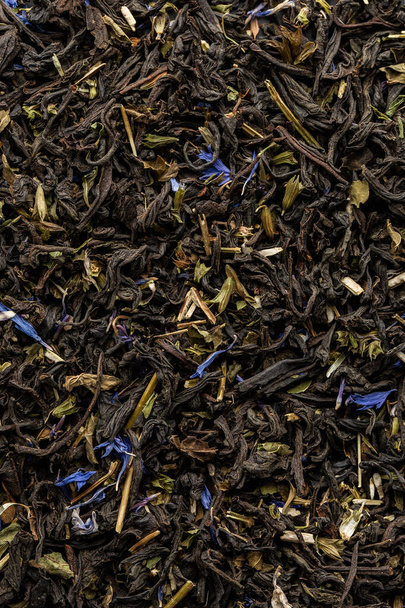 Heap of dry tea leaves. Dried black tea. Textured background of dried leaves of aromatic black tea with blue flowers. - Фото, изображение