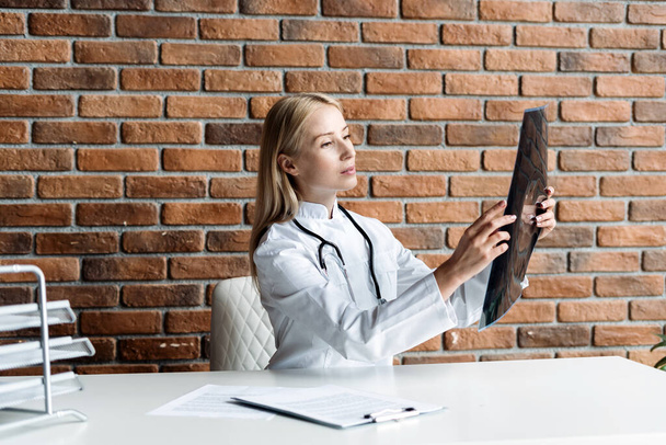 Concentrated and serious physician looking at x ray picture, diagnoses a patient injury. Doctor wearing in white coat uniform with stethoscope sitting in office at private clinic, checking mri scan - Foto, Bild