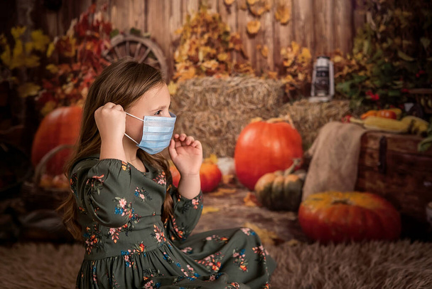 Portrait of a girl in a witch costume, preparing for Halloween. Puts on a medical face mask that protects against the virus. Dark autumn background with pumpkins - Photo, Image