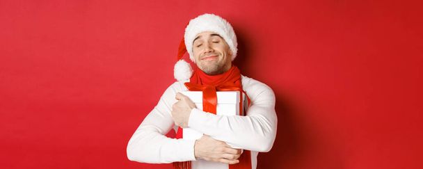 Concept de vacances d'hiver, Noël et style de vie. Image d'un charmant gars en chapeau et écharpe de Père Noël, étreignant son cadeau de nouvelle année et souriant flatté, debout sur fond rouge - Photo, image