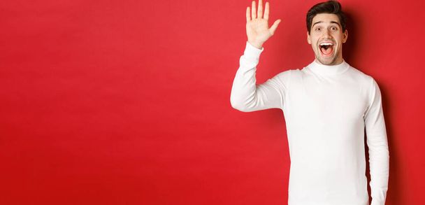 Obrázek šťastného a přátelského mladíka, jak pozdravuje, mává rukou, aby někoho pozdravil, stojí nad červeným pozadím - Fotografie, Obrázek
