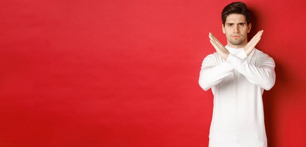 Изображение серьезного и злого человека в белом свитере, выражающего неодобрение хранения, показывающего поперечный знак, чтобы остановить что-то плохое, запретить действие, стоя на красном фоне - Фото, изображение