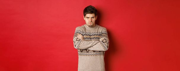 Képek a dühös és offedned srác karácsonyi pulóver, érzés őrült, keresztbe karok a mellkason és duzzogás, álló felett piros háttér - Fotó, kép