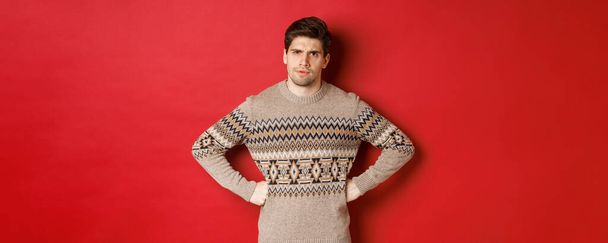 Retrato de hombre dudoso y decepcionado, vistiendo suéter de Navidad, frunciendo el ceño y mirando disgustado a usted, de pie sobre fondo rojo - Foto, imagen