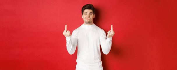 Image of pissed-off and distressed man telling to fuck off, showing middle-fingers and looking upset, standing over red background in white sweater - Fotó, kép