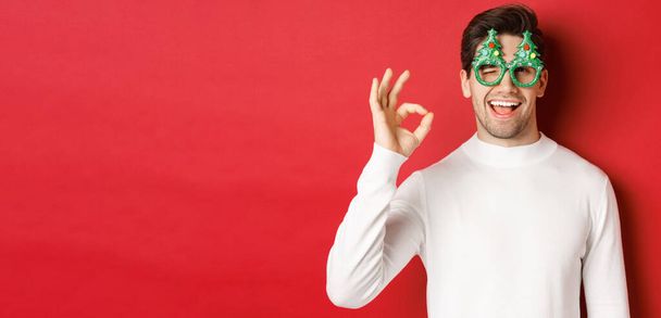 Arckép jóképű és pimasz fickó, visel party szemüveg és fehér pulóver, mutatja rendben jel és kacsintás, kívánva boldog új évet, áll a piros háttér felett - Fotó, kép