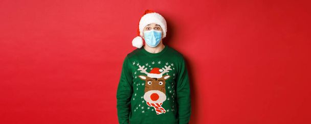 Koncepcja nowego roku, covid-19 i dystans społeczny. Zaskoczony gość w czapce Mikołaja, masce medycznej i swetrze świątecznym, wyglądający na zachwyconego kamerą, stojący na czerwonym tle - Zdjęcie, obraz