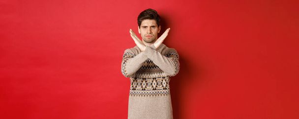 Imagen de hombre guapo enojado y serio en suéter de Navidad, diciendo no o parar, mostrando gesto cruzado para restringir algo, prohibir la acción, de pie sobre el fondo rojo - Foto, imagen