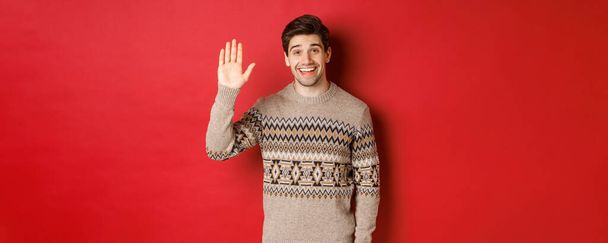 Портрет красивого и дружелюбного парня в рождественском свитере, здоровающегося и машущего рукой, приветствующего кого-то и желающего счастливых зимних праздников, стоящего на красном фоне - Фото, изображение