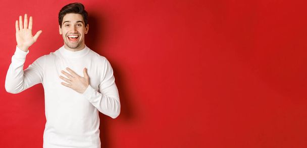 Portrait of honest smiling man in white sweater, making a promise, swearing to tell truth, standing against red background - Photo, Image