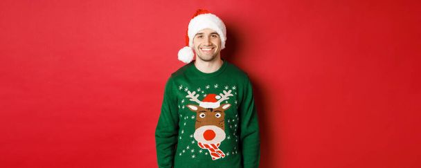 Pojęcie ferii zimowych, Bożego Narodzenia i stylu życia. Przystojny facet z włosia, w kapeluszu Mikołaja i zielonym swetrze, uśmiechnięty radosny, świętujący Nowy Rok, stojący na czerwonym tle - Zdjęcie, obraz